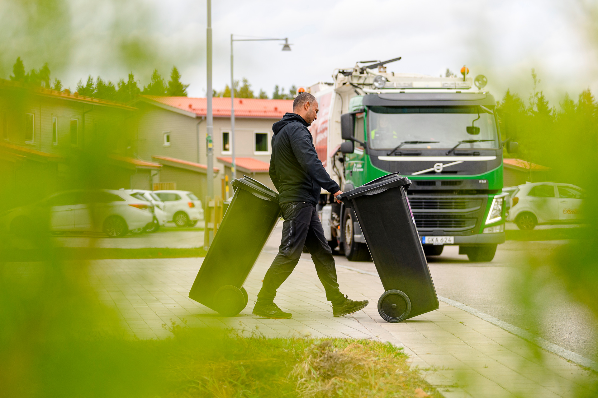 Man drar ett kärl framför sig och ett kärl bakom sig och fronten på en sopbil syns i bakgrunden