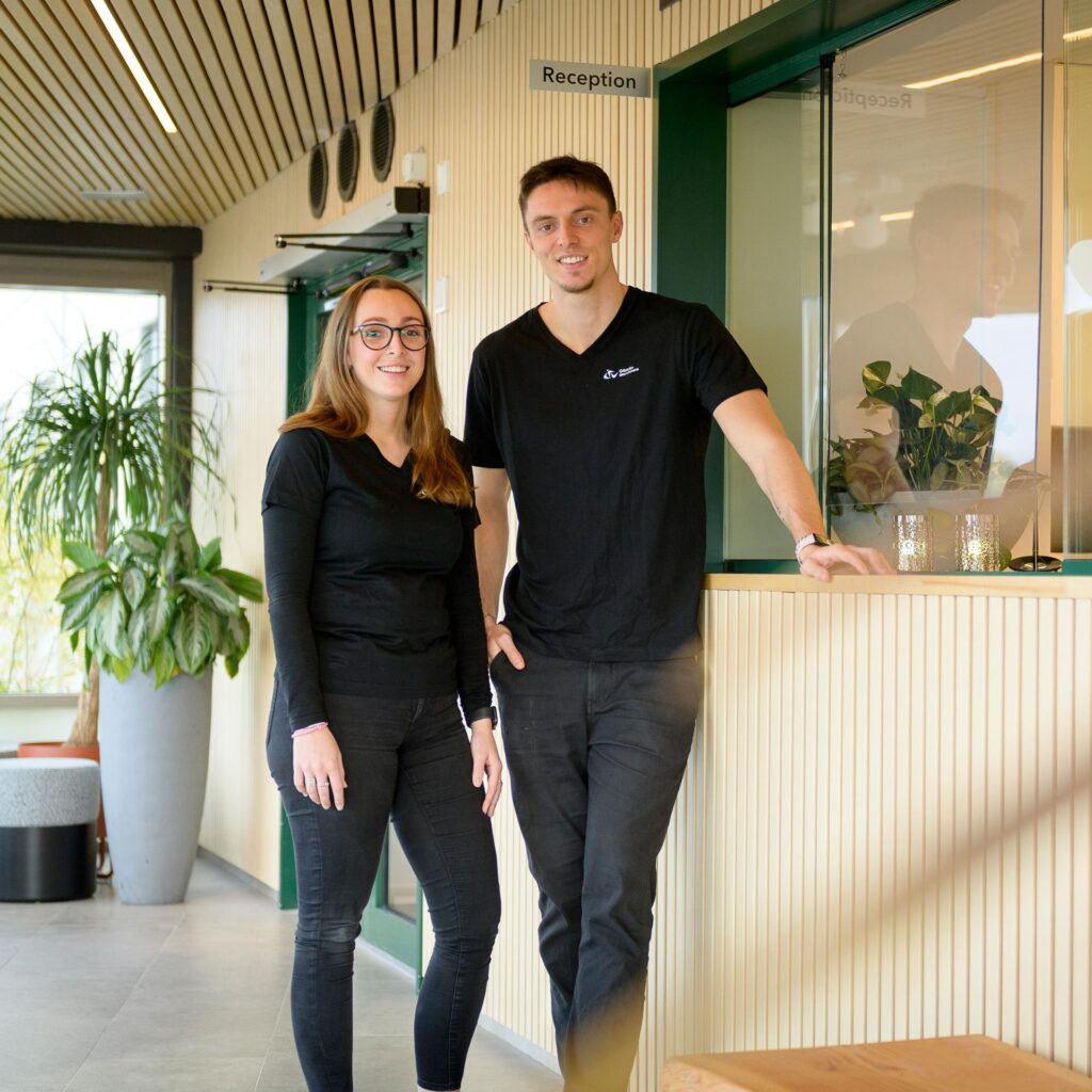 En kvinna och en man står framför en reception med träpanel, grön växt i bakgrunden