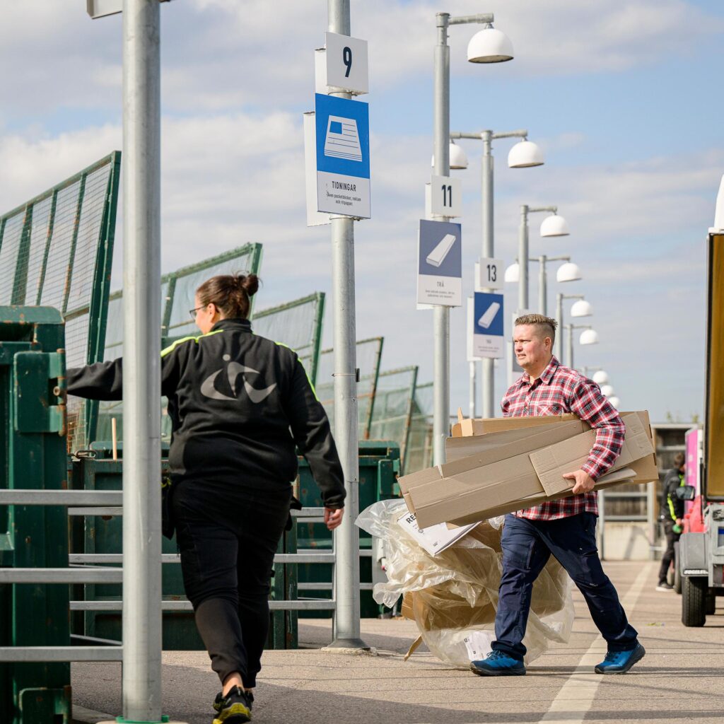 man med förpackningar under armen går mot container, hänvisad av personal