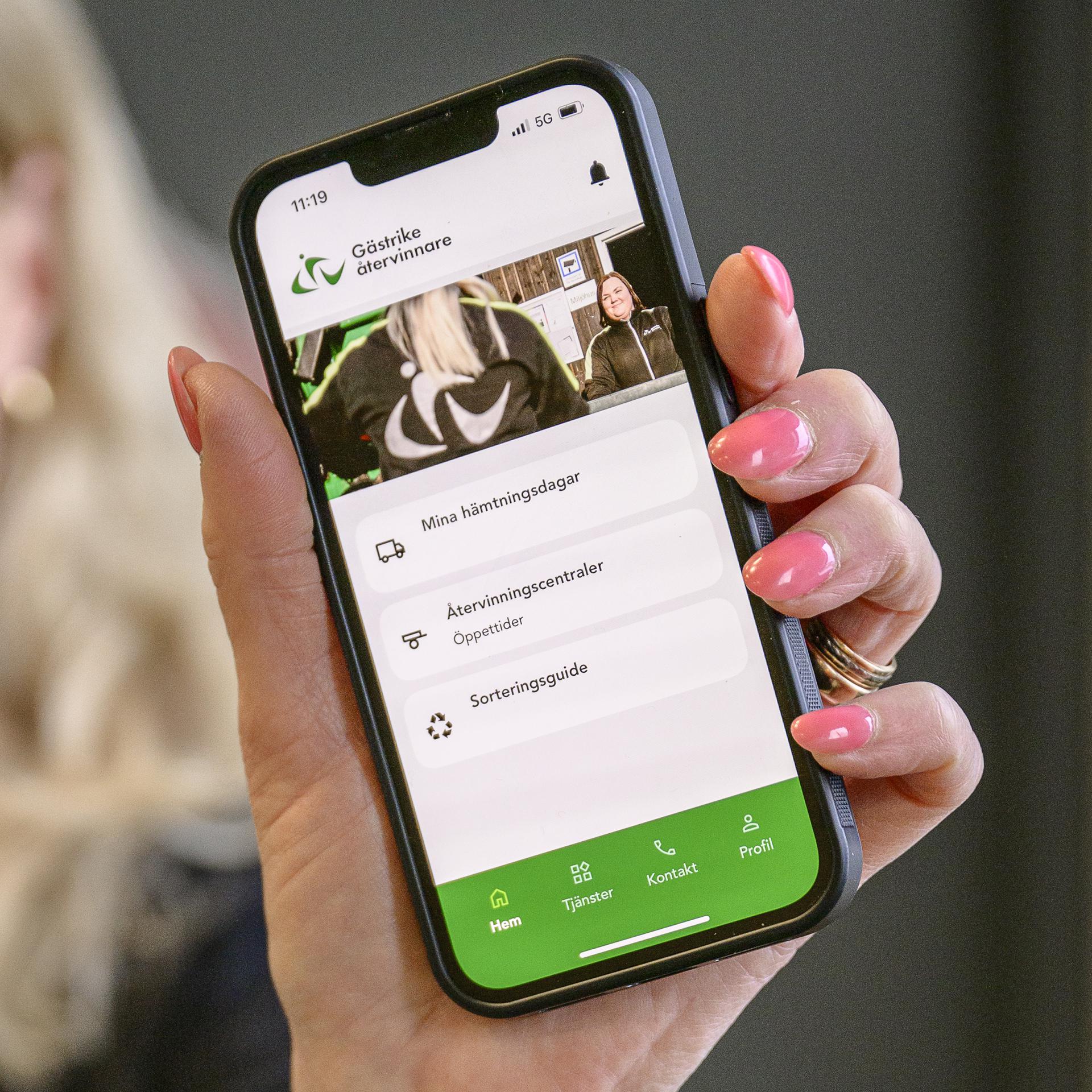 En hand håller fram en mobiltelefon som visar Gästrike återvinnares app